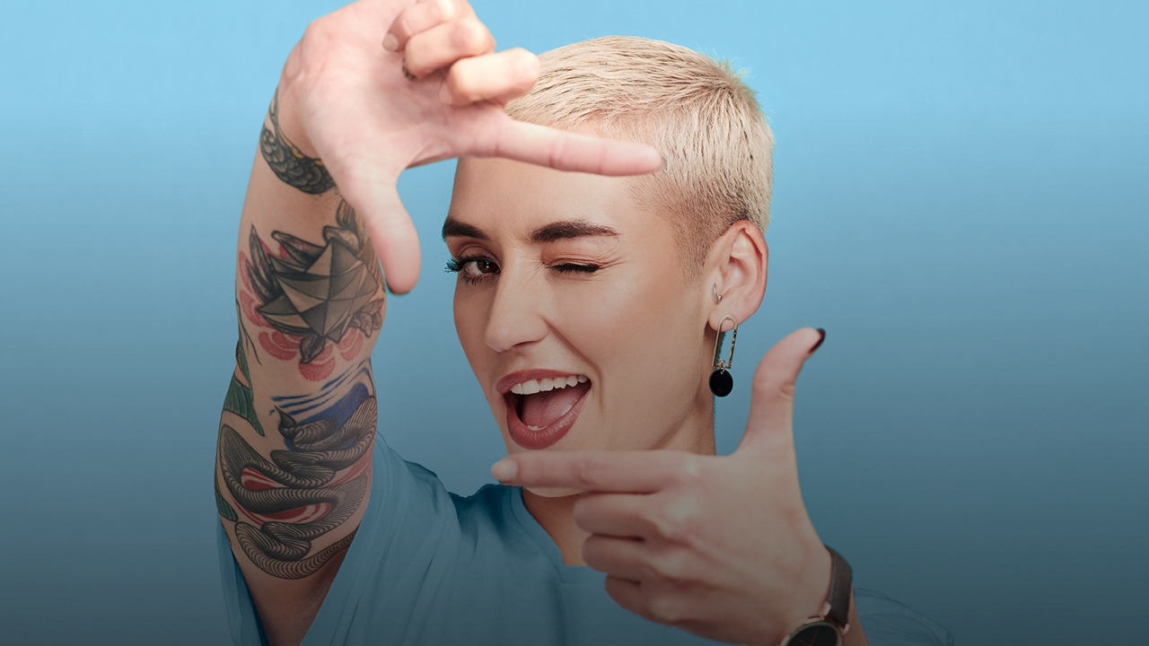 Eine Frau mit kurzem Haar formt mit ihren Händen ein Viereck und blickt lachend dadurch hindurch in die Kamera.